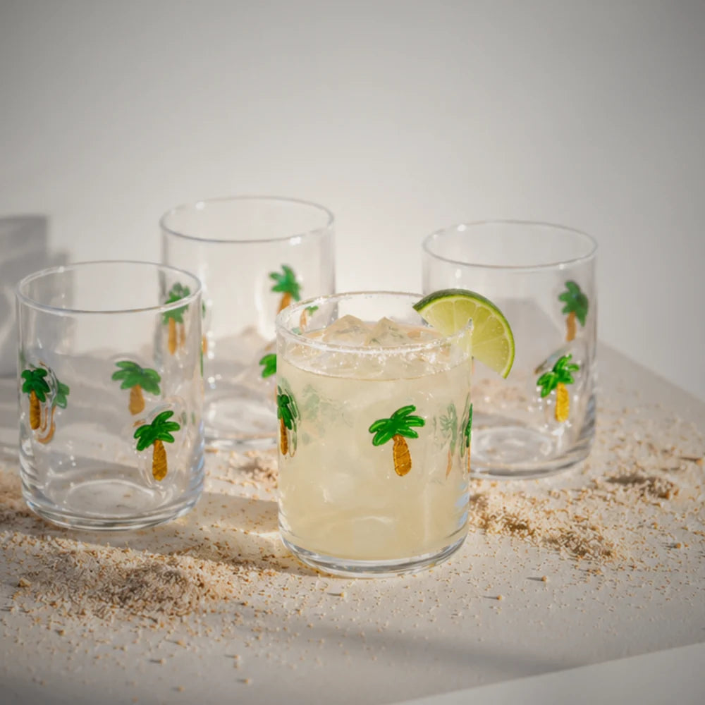 Clear glass tumblers with a palm tree print, one holding a drink with a lime wedge, on sand