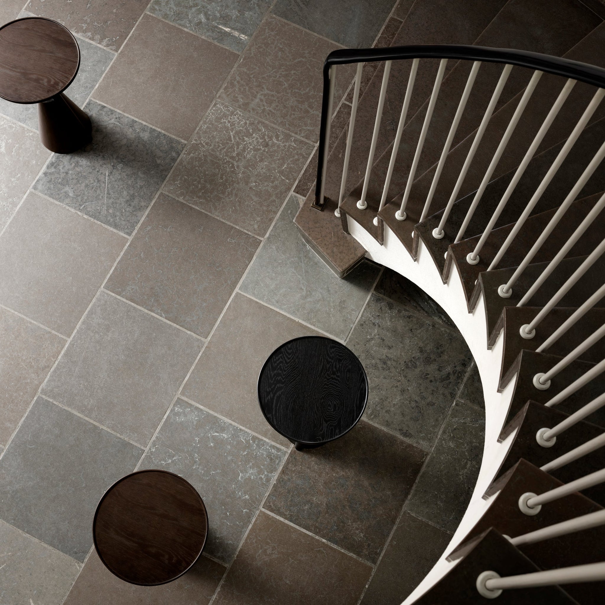 Modern side tables with white frame on a dark tiled floor, beneath a winding stair case. 