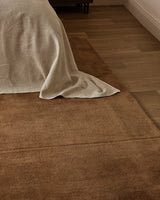 Bastion rug in hazelnut at the foot of a bed with oatmeal linen bedding, on oak flooring